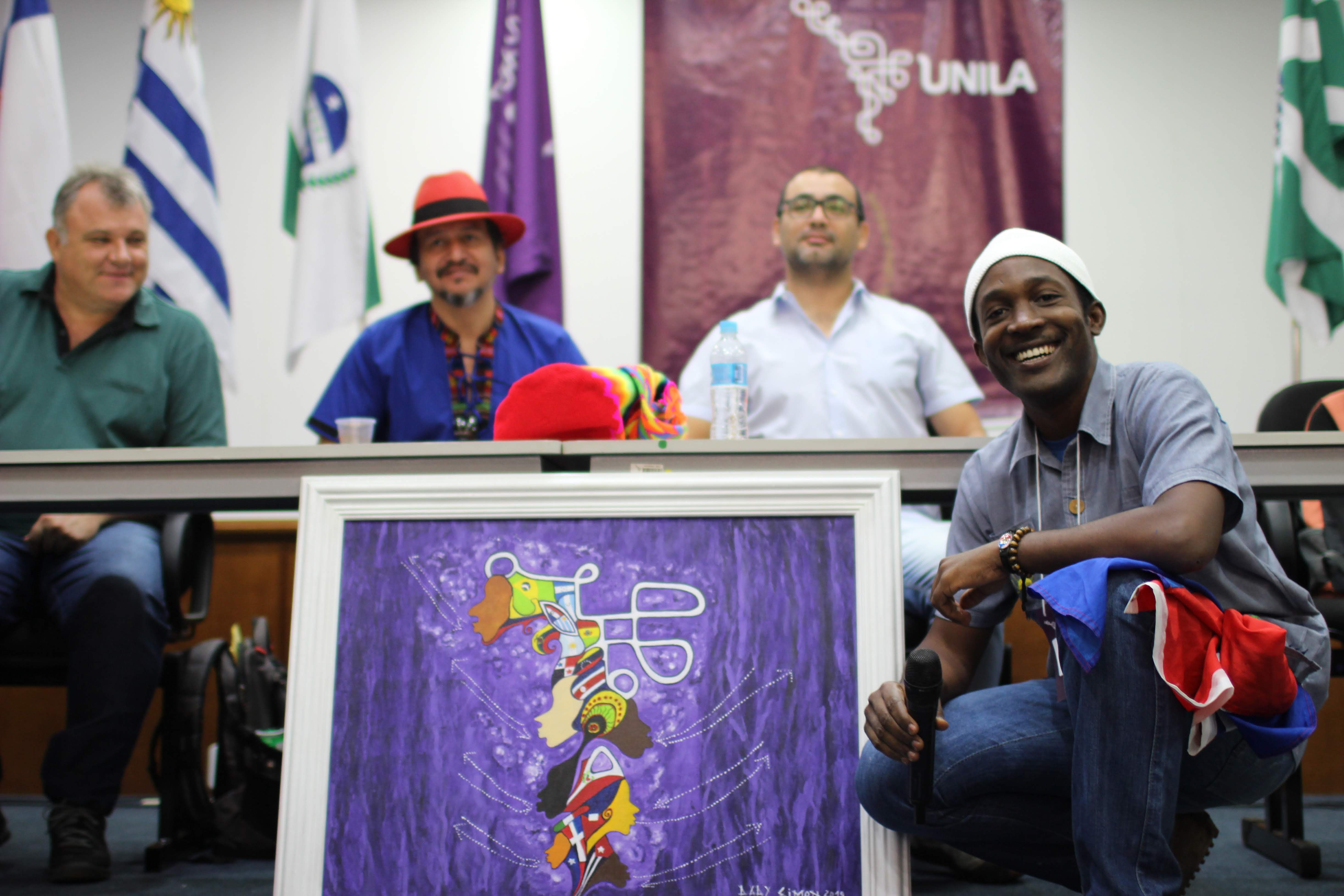 Dady Simon, artista haitiano, com quadro pintado por ele à frente da mesa onde estão coordenadores de cursos da UNILA
