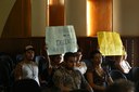 Estudantes sentados no auditório seguram cartazes pedindo mais professores para o curso de Administração Pública 