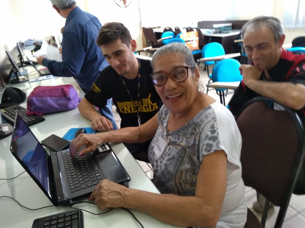 Senhora idosa utiliza um computador e é observada por um estudante que a orienta