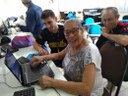 Senhora idosa utiliza um computador e é observada por um estudante que a orienta